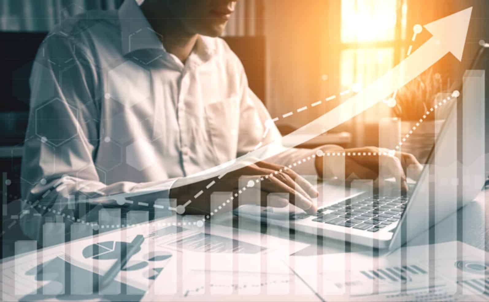 Business man on his laptop on a desk with paper graphs spread out and virtual arrow pointing up for growth